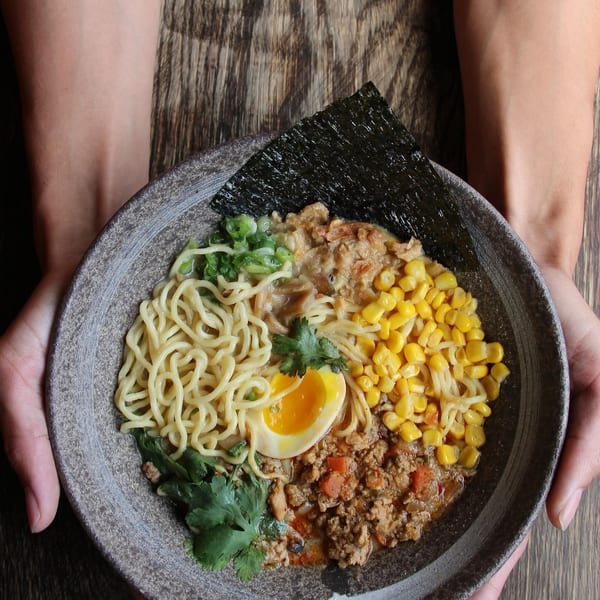 Hands holding a bowl of ramen with noodles, corn, ground meat, a soft-boiled egg, and nori.