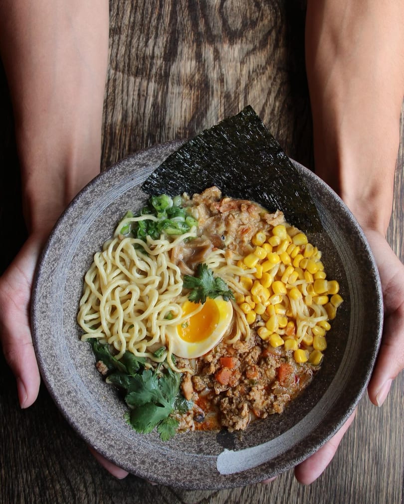 Hands holding a bowl of ramen with noodles, corn, ground meat, a soft-boiled egg, and nori.