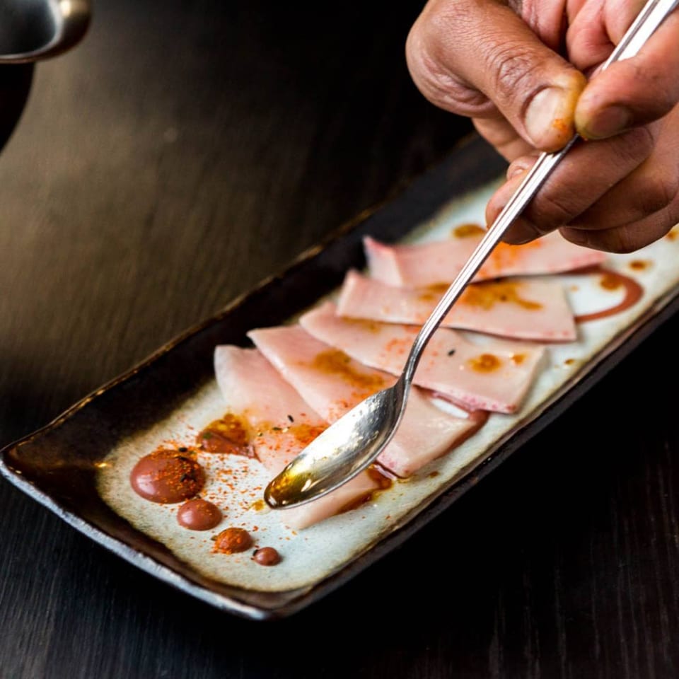 A chef's hand drizzles sauce onto thinly sliced fish on a rectangular plate.