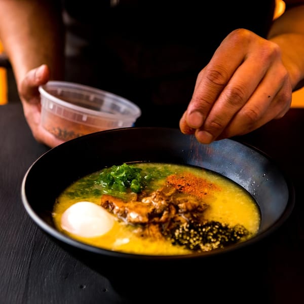 A chef's hands sprinkle spices over a vibrant bowl of ramen with egg and green onions.