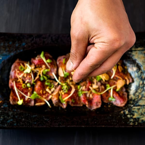 A hand sprinkles garnish over thinly sliced beef tataki on a dark rectangular plate.
